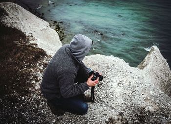 High angle view of man with camera crouching on cliff by sea