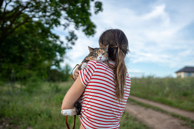 Woman pet owner holding and hugging domestic cat in hands walking outside among countryside
