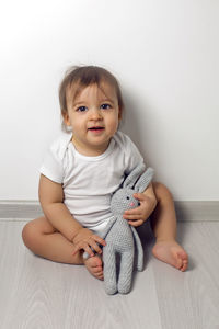 Baby boy in white clothes sitting on the floor with a gray knitted rabbit at the white wall 