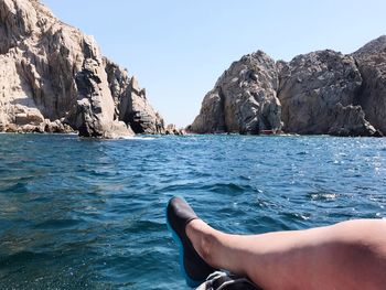 Low section of woman wearing shoe against sea and rock formations during sunny day