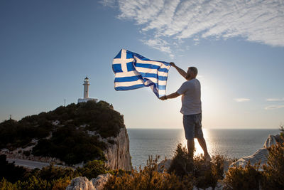 Rear view of man with arms outstretched standing by sea against sky