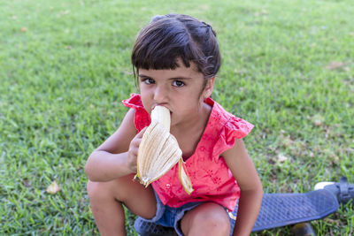 Little girl eating banana sitting outdoors on the grass in a park.