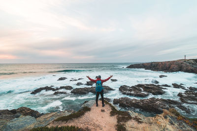 Rear view of woman standing on rock at beach against sky during sunset