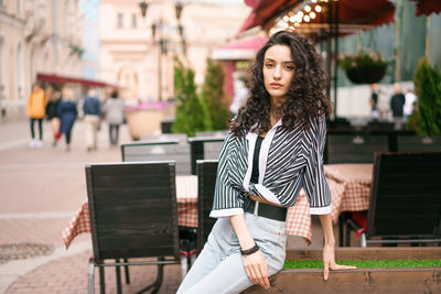  woman  the street at the cafe . dark curly hair brown eyes .there are tables and chairs in the back
