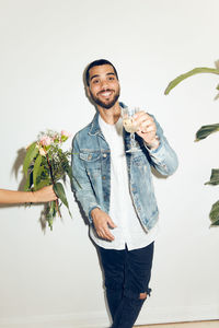 Cropped hand of woman giving flowers to smiling young man holding with drink while standing against white wall at home