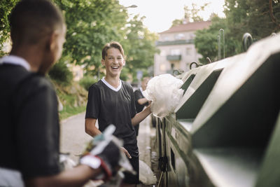 Happy teenage boy putting plastic garbage bag in recycling bin near friends