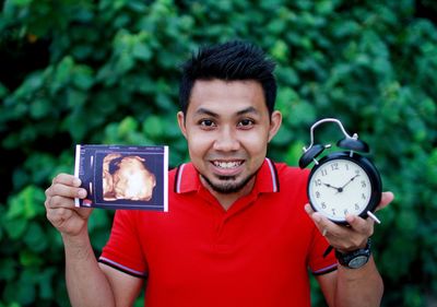 Portrait of young man holding alarm clock with ultrasound standing against plants