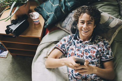 High angle portrait of happy teenage boy listening music through smart phone on bed at home