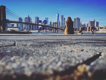 Rear view of woman sitting by brooklyn bridge and city skyline against clear blue sky