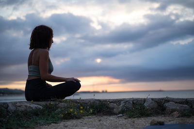 Side view of woman sitting on field against sky during sunset