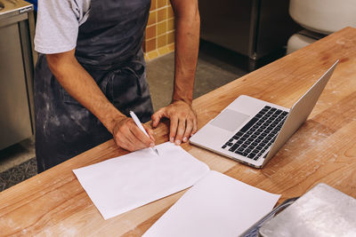 Cropped unrecognizable male bakery owner standing at counter writing notes on paper and using laptop and ingredients for baking working