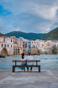 Rear view of woman sitting on bench against buildings in city