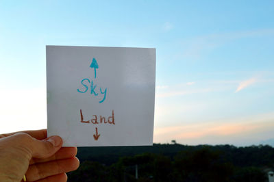 Close-up of hand holding paper with text against sky