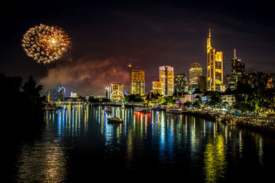 Firework display over river by illuminated buildings against sky