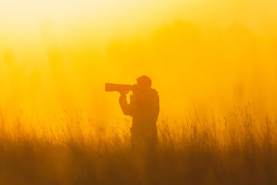 Side view of man photographing against orange sky