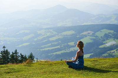 Young woman meditating on grassy field against mountains during foggy weather