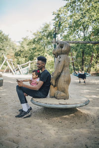 Young man using mobile phone while sitting with daughter on outdoor play equipment