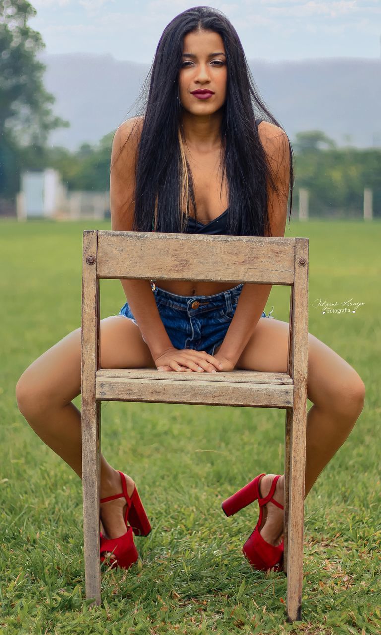 sitting, one person, long hair, hairstyle, grass, young adult, women, adult, portrait, full length, seat, brown hair, plant, relaxation, nature, fashion, front view, looking at camera, clothing, chair, leisure activity, casual clothing, person, limb, lifestyles, field, human leg, outdoors, looking, green, sky, emotion, summer, contemplation, female, land, day, park, shoe, smiling, black hair, meadow, plain, teenager, park - man made space, bench, landscape, dress, serious