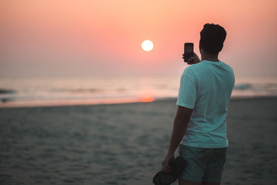 Rear view of man standing at beach against sky during sunset