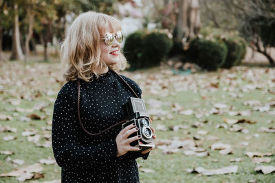 Smiling young woman holding vintage camera while standing on land during autumn