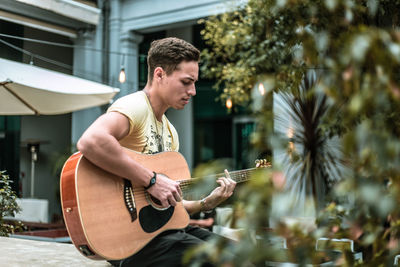 Young man playing guitar while sitting against building