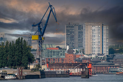 Cranes at harbor against buildings in city against sky