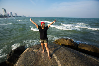 Young woman arms outstretched standing on rock at seashore against sky