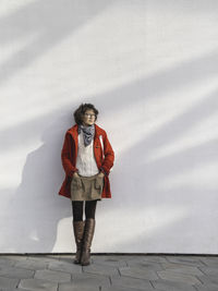 Curly woman in red duffle coat is standing by white wall with sunbeams. smiling student. 