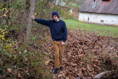 Handsome young man leans on tree on autumn hiking trail by idyllic stone cottage