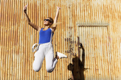 Full length of young woman jumping against wooden wall