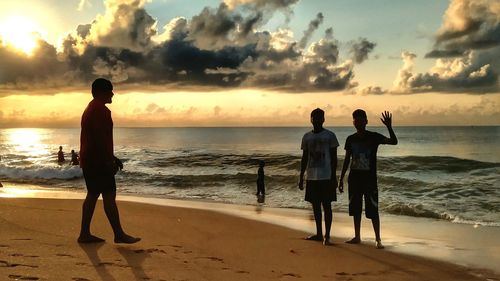 Rear view of men standing on beach against sky during sunset