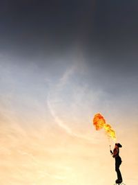 Low angle view of shirtless man blowing fire while standing against orange sky