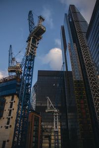 Low angle view of cranes by skyscrapers in city against sky
