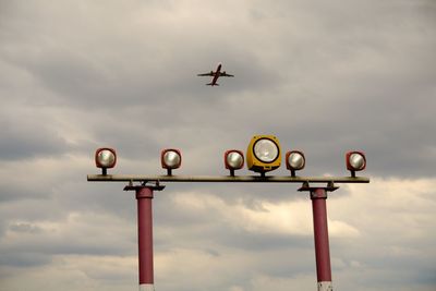 Low angle view of airplane flying over lighting equipment against cloudy sky