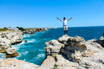 Full length of man with arms outstretched standing on rock by sea against clear sky