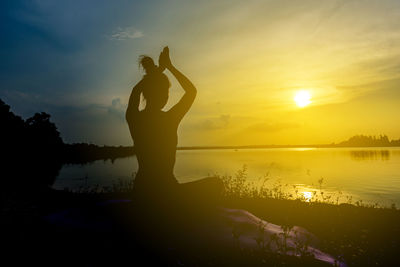 Silhouette woman exercising at lakeshore against sky during sunset