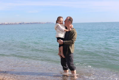 Father carrying daughter while standing in sea at beach against sky