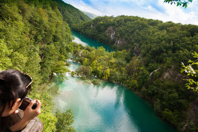Man photographing lake by trees against sky