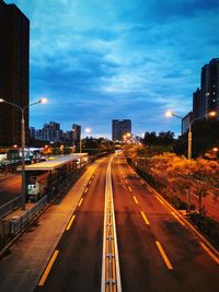Illuminated road by buildings against sky at dusk