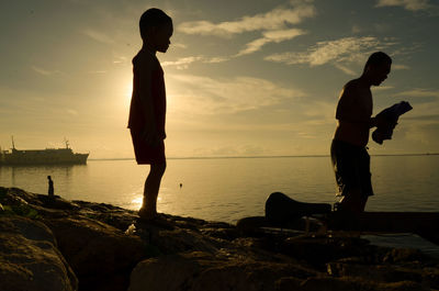 Silhouette boy standing on rock by sea against sky during sunset