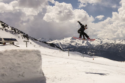 Full length of snowboarder jumping against cloudy sky at snowcapped mountain