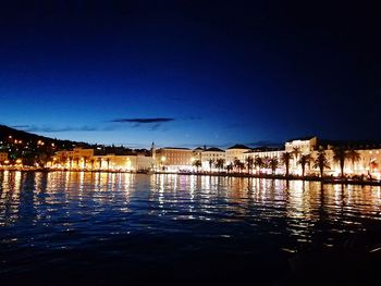 Illuminated buildings by river against blue sky at night