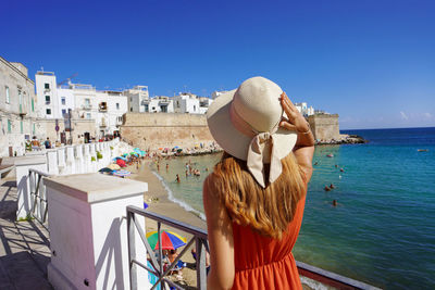 Back view of young woman with hat and dress enjoying seascape in monopoli town, puglia, italy.
