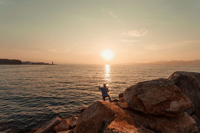 Man standing on rocks by sea against sky during sunset