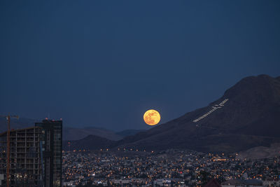 Aerial view of moon over illuminated city against clear sky at dusk