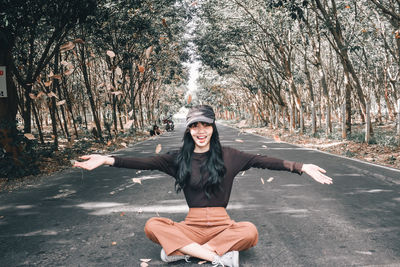 Portrait of smiling young woman throwing leaves while sitting on road amidst trees in forest