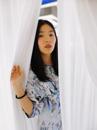 Portrait of young woman standing by curtain at home