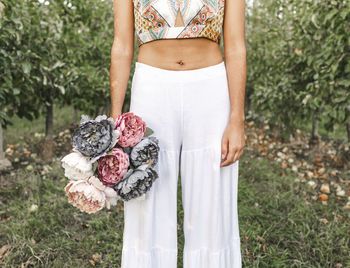 Crop unrecognizable female with bouquet of blooming peonies standing in garden with rows of apple trees in farm