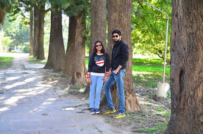 Portrait of couple standing by tree trunk on footpath in park