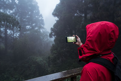 Side view of anonymous hiker in hoody raincoat standing on footbridge and taking picture of nature while hiking alone in green coniferous forest in foggy rainy weather